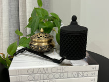 Load image into Gallery viewer, A matte black medium soy candle with a geometric pattern jar, placed on a stack of books, with a plant in the background.