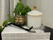 Load image into Gallery viewer, A white soy candle with a geometric pattern jar and a gold-colored lid, placed on a stack of books with a plant in the background.