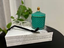 Load image into Gallery viewer, A medium-sized soy candle with a geometric pattern jar, lid off, placed on a stack of books next to a plant. The candle is teal in color.