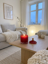 Load image into Gallery viewer, Essence red glass soy candle. Cozy living room with a red candle on a wooden coffee table, soft lighting, and a window in the background.
