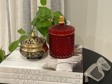 Load image into Gallery viewer, A red soy wax candle in a medium-sized geometric cut glass jar with a gold lid, placed on a surface next to a plant and decorative items.
